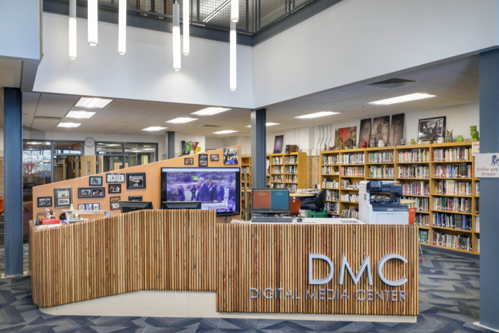 The reception desk of the Digital Media Center in a library, featuring a slatted wood facade, computer monitors, and bookshelves in the background.