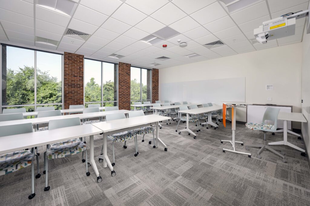 A modern classroom featuring rows of tables with chairs, a whiteboard on the wall, and large windows with a view of trees.