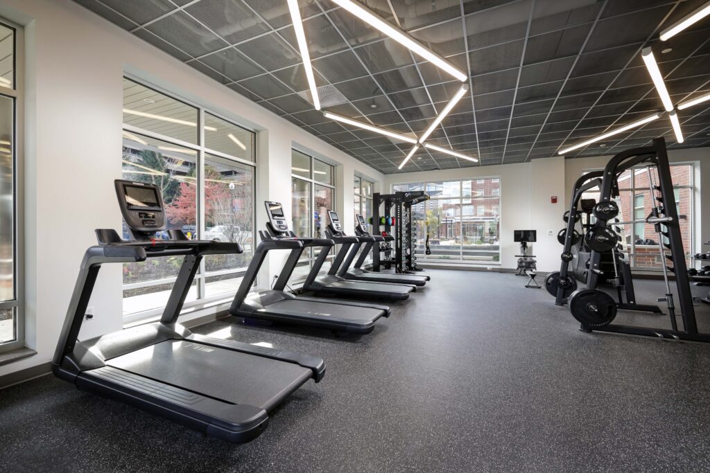 A fitness center with a row of treadmills and a weightlifting station near large windows.