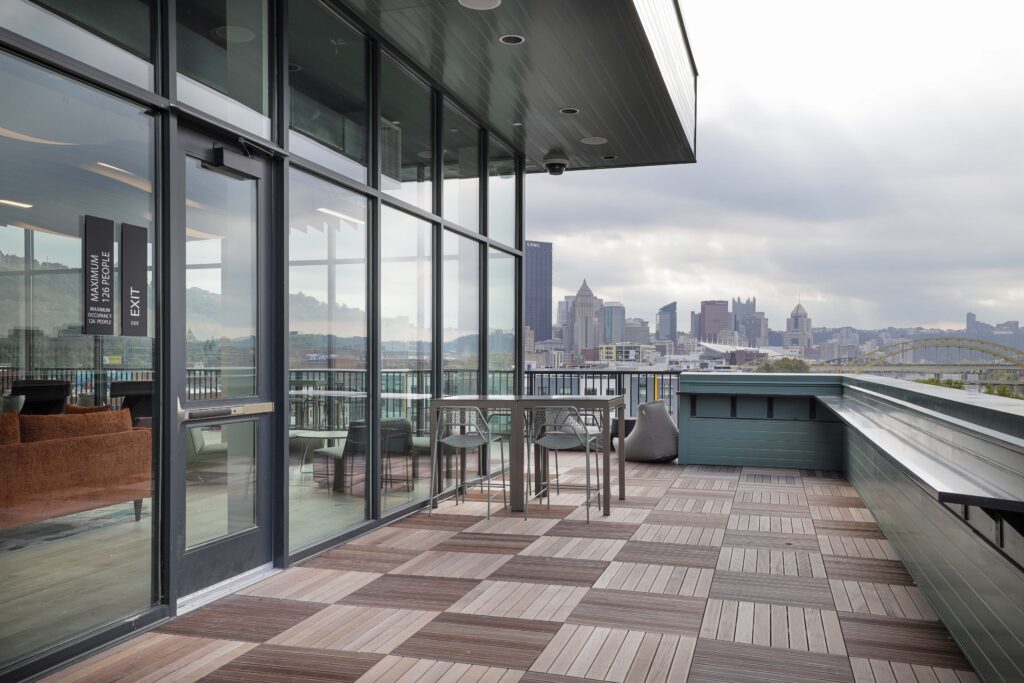A rooftop terrace featuring a glass-walled indoor area, outdoor seating, and a view of the Pittsburgh skyline.