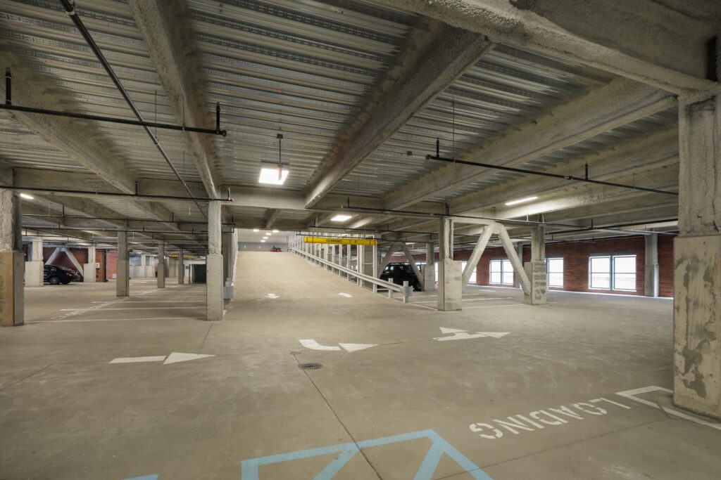 An indoor parking garage with concrete pillars, a central vehicle ramp, and painted directional arrows on the floor.