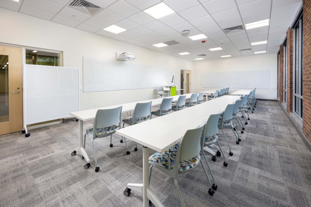 A classroom with rows of white folding tables and patterned chairs facing a long wall-mounted whiteboard and projector.