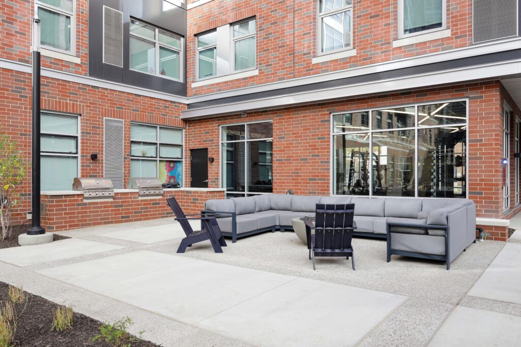 Outdoor patio with a large grey sectional sofa, two dark chairs, a fire pit, and two built-in stainless steel grills against a red brick building.
