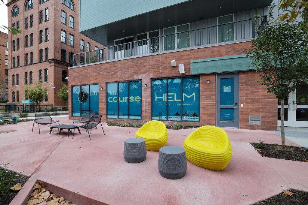 Exterior of The Helm on the Allegheny building, featuring brick and teal-accented architecture, glass signage, and outdoor seating on a paved patio.