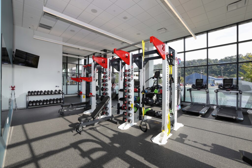 Fitness center with a central strength training rig, dumbbell racks, and treadmills along a windowed wall.