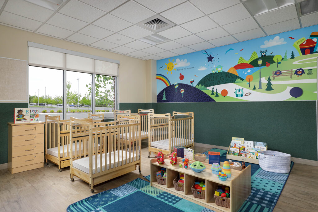 A childcare room featuring several wooden cribs, a low shelf with toys and books, and a colorful mural of a landscape on the wall.