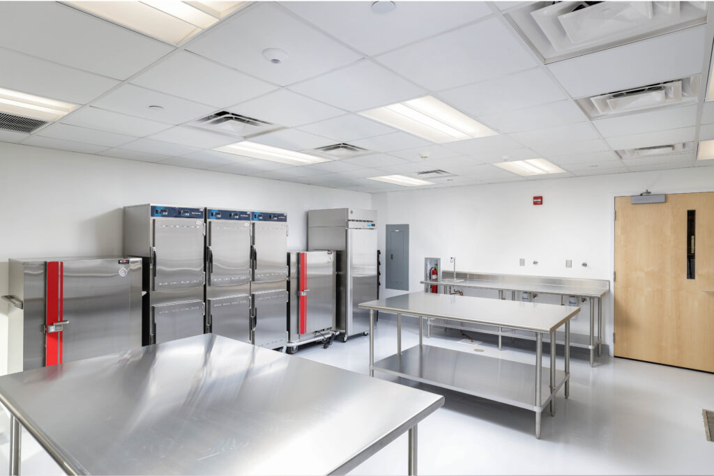 A commercial kitchen featuring stainless steel prep tables, multiple tall refrigerators, and a stainless steel sink along the wall.