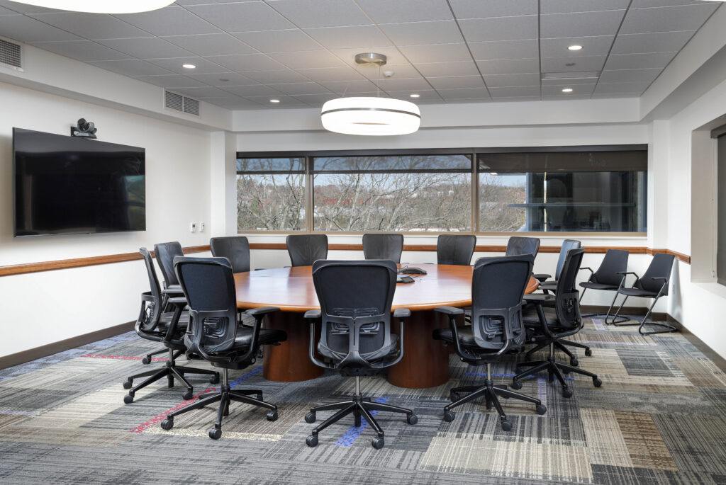 A round conference table surrounded by black ergonomic chairs in a room with a wall-mounted monitor and large windows.