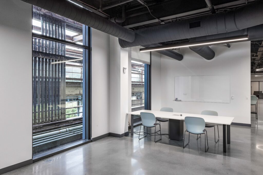 A modern meeting room with a long white table, four blue chairs, and a whiteboard, featuring floor-to-ceiling windows overlooking an industrial exterior.