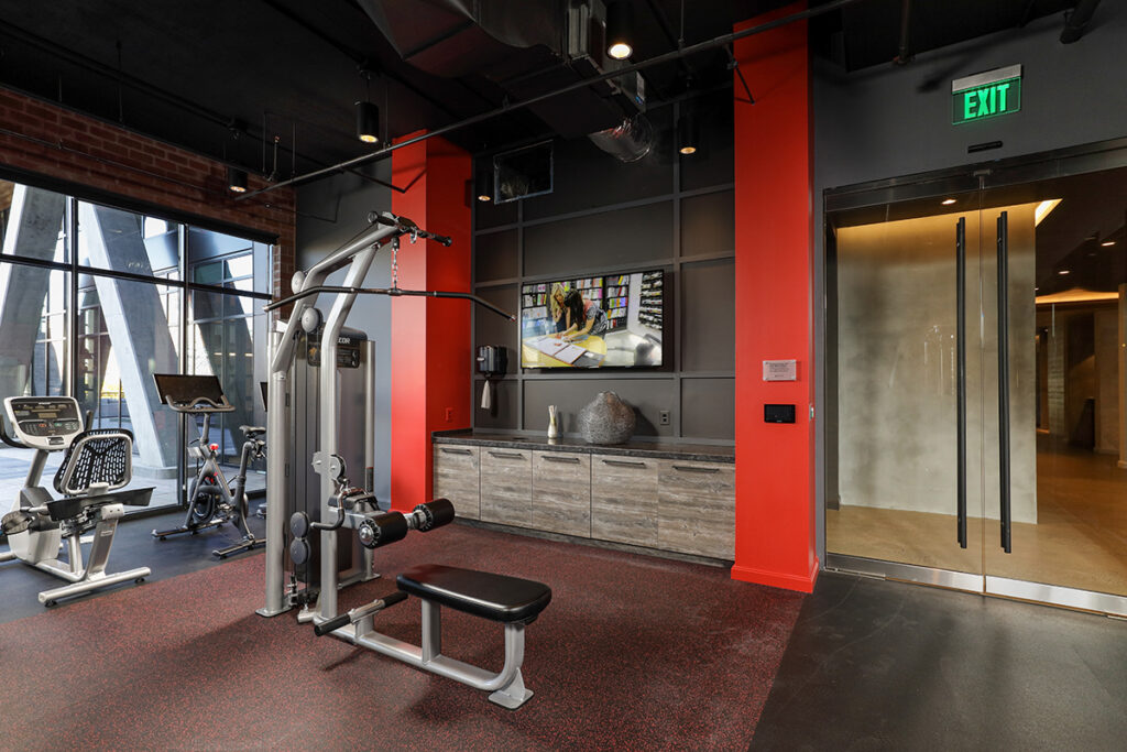 Gym with a lat pulldown machine, stationary bikes, a wall-mounted television, and a glass door with an exit sign.