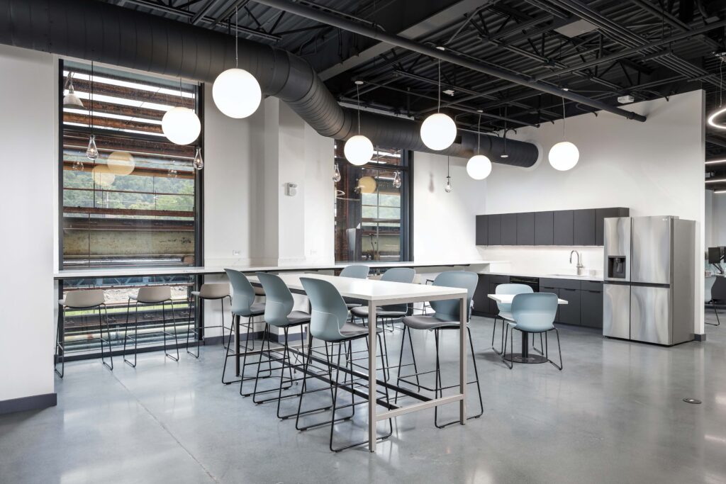 Office break room with a high communal table, stools, and a kitchenette featuring dark cabinets and a stainless steel refrigerator.