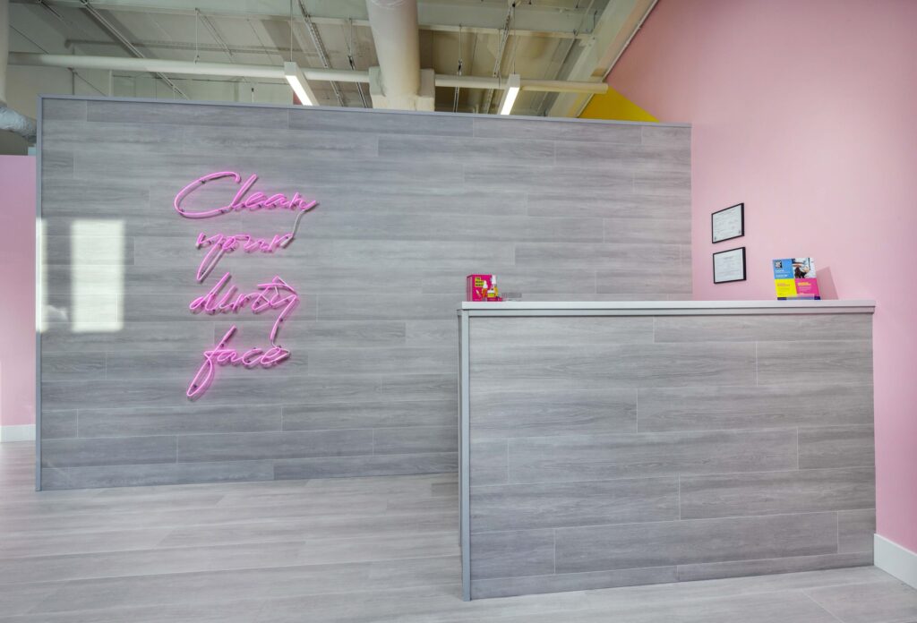 Reception desk with a grey wood-paneled wall featuring a pink neon sign that reads “Clean your dirty face.”