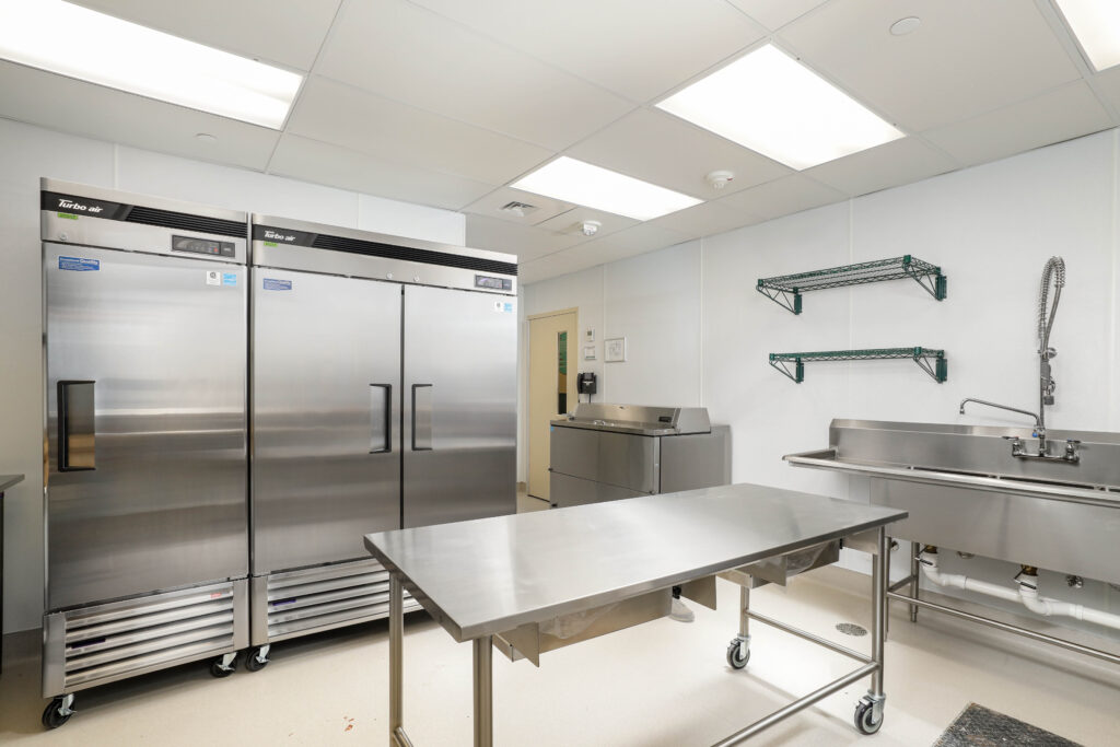 A commercial kitchen with two stainless steel refrigerators, a central work table, a sink, and wall-mounted shelves.