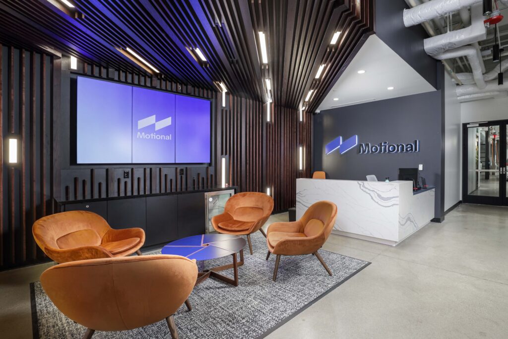 Lobby area featuring the Motional logo on a screen and a reception desk, with four orange lounge chairs arranged around a circular table.
