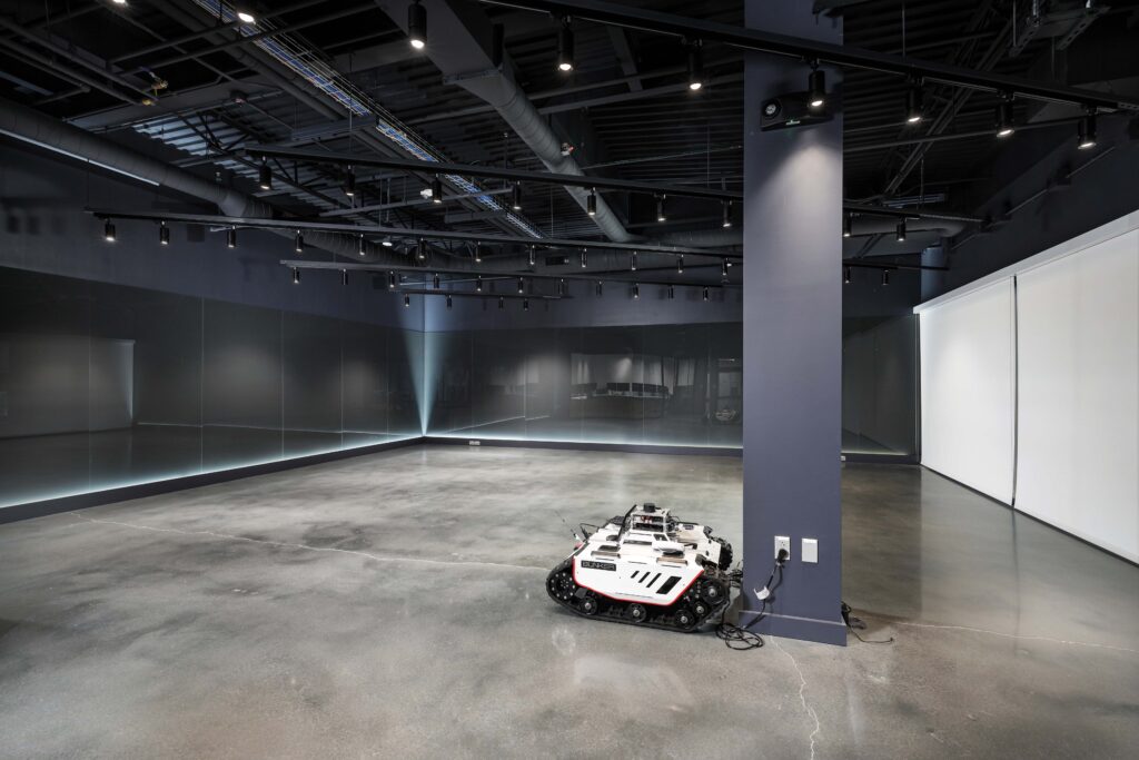 A white, tracked autonomous robot plugged into an outlet in a large, empty commercial space with dark walls and polished concrete floors.