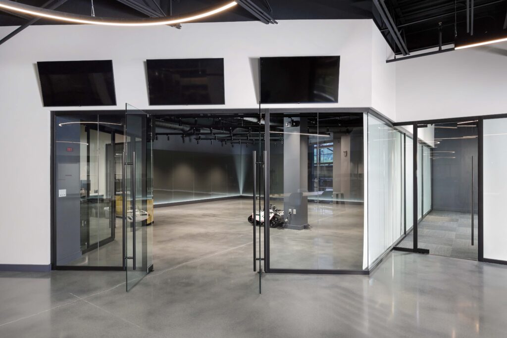 Modern office interior with glass-walled conference rooms, concrete floors, and three flat-screen monitors mounted on the wall.
