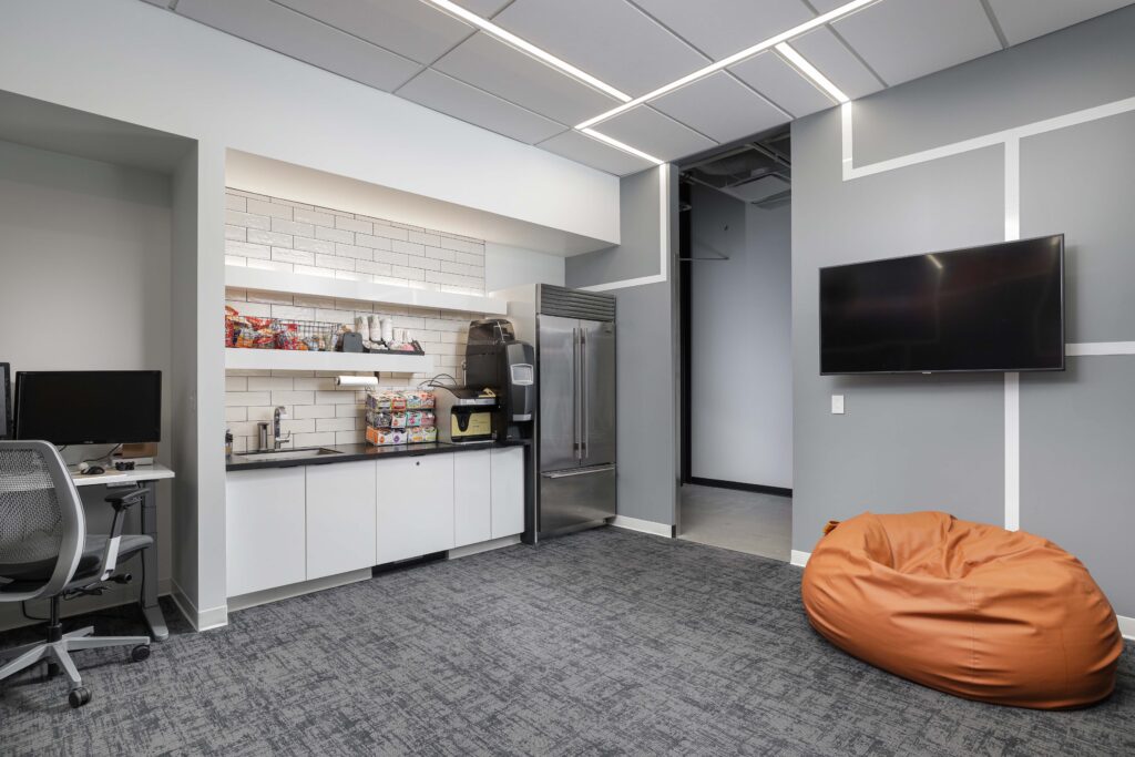 An office break room with a kitchenette, stainless steel refrigerator, coffee maker, wall-mounted television, and an orange bean bag chair.
