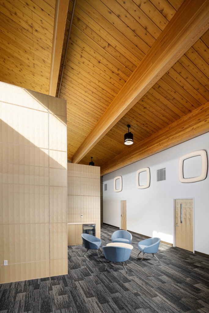 A modern lobby with light wood paneling, a vaulted wood ceiling, and four blue armchairs arranged around a small round table.