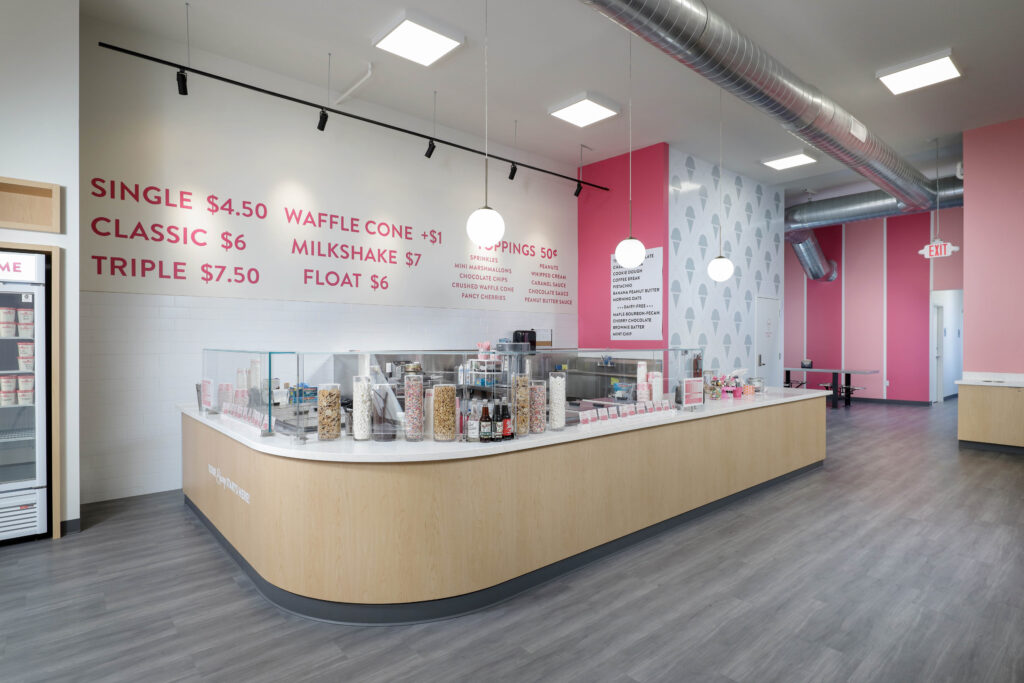 Ice cream shop interior with a curved service counter, glass topping dispensers, and menu pricing written on the wall.