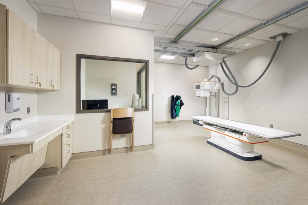 A radiology room featuring an X-ray table, a ceiling-mounted imaging unit, a sink, and an observation window.