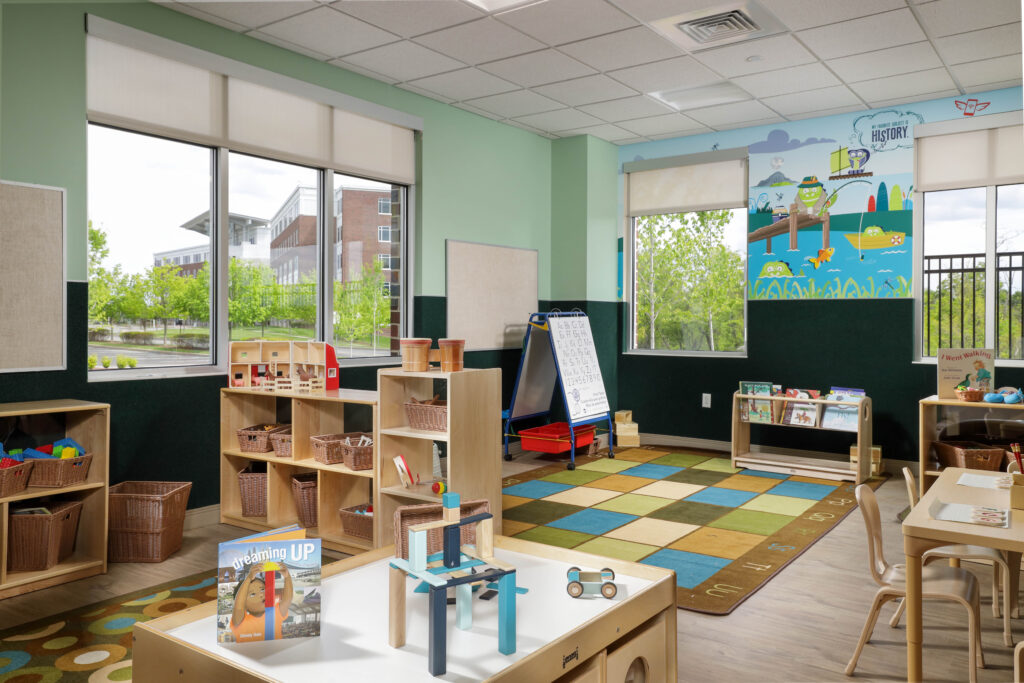 A preschool classroom with wooden shelves, a play table, a colorful rug, and a whiteboard easel.