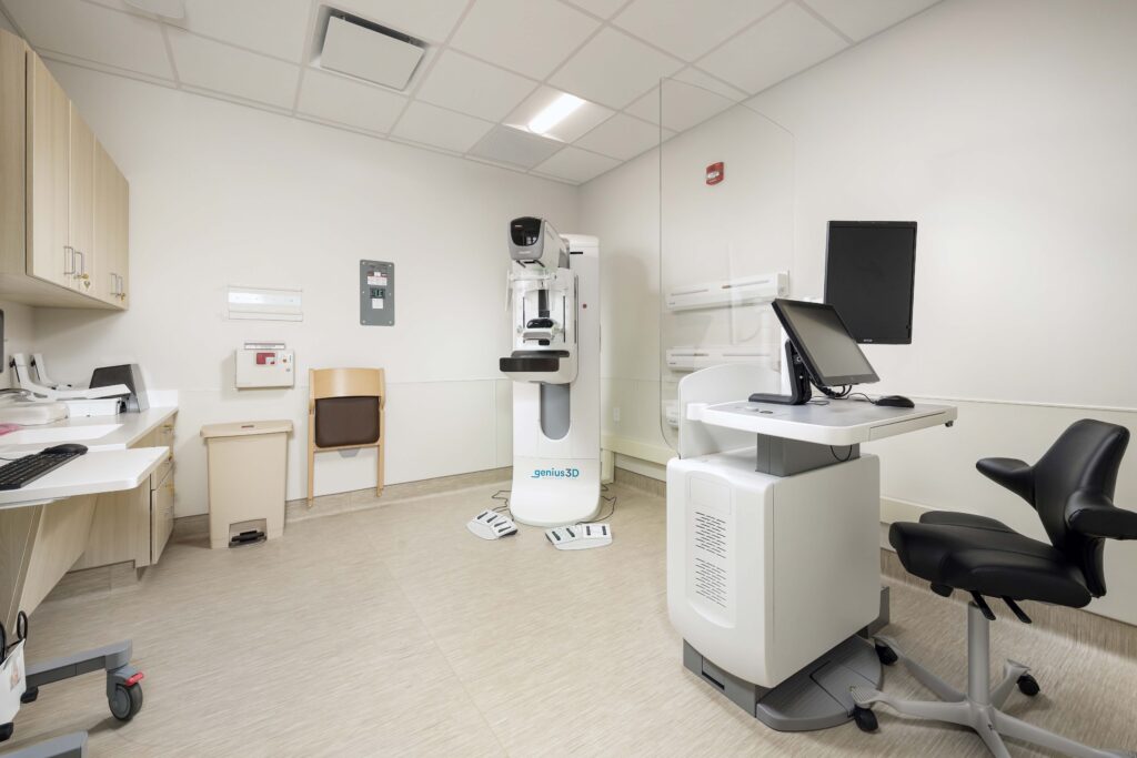 A Genius 3D mammography machine in a medical exam room with an associated computer workstation.