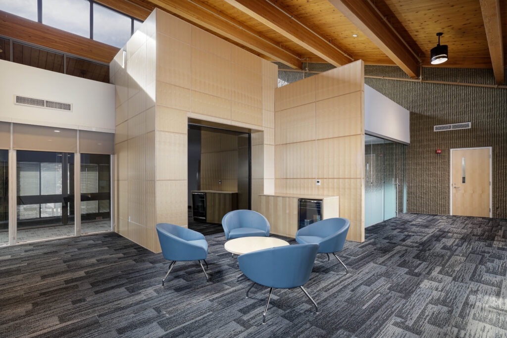 A small lounge area with four blue armchairs around a circular table, set within an office space featuring light wood paneling, exposed ceiling beams, and gray carpet.