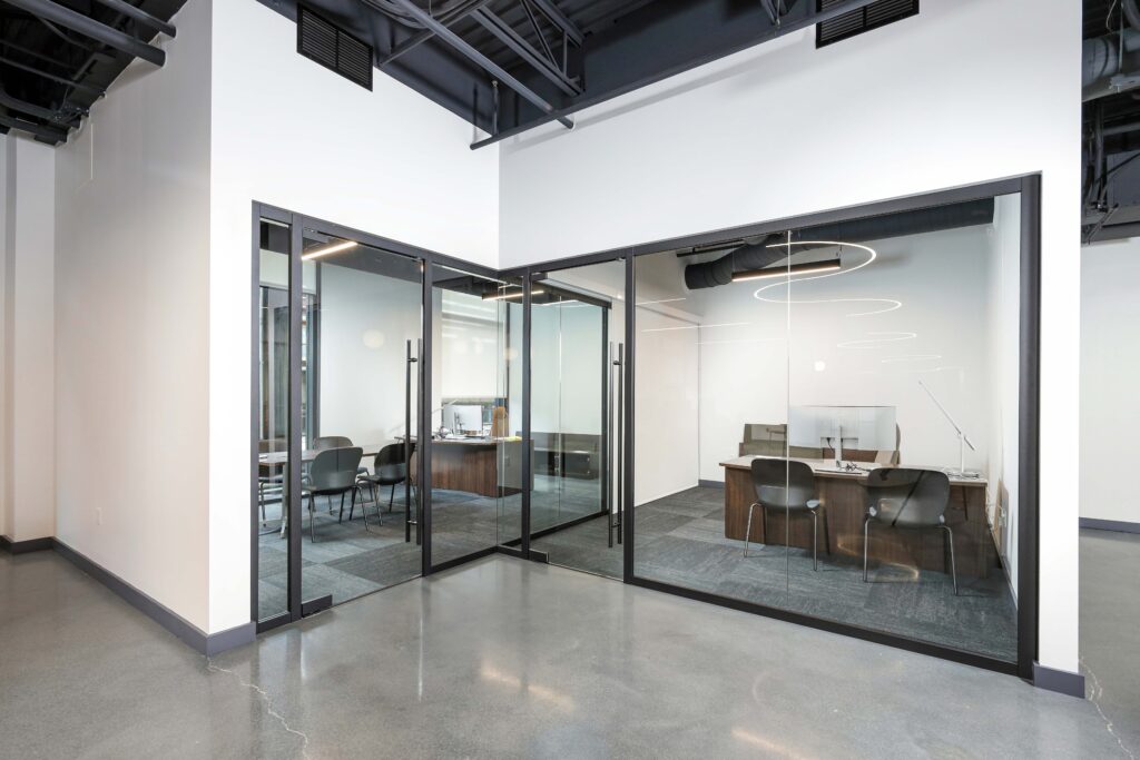 Modern office space with glass-walled conference rooms containing desks, chairs, and computers on a polished concrete floor.