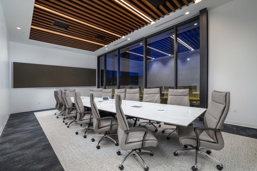 A modern boardroom with a long white table, grey leather office chairs, and a slatted wood ceiling.