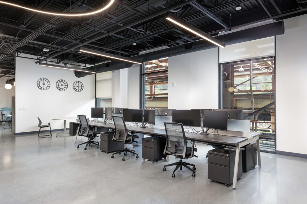 Modern open-plan office with rows of computer desks, rolling chairs, and three large wall clocks.