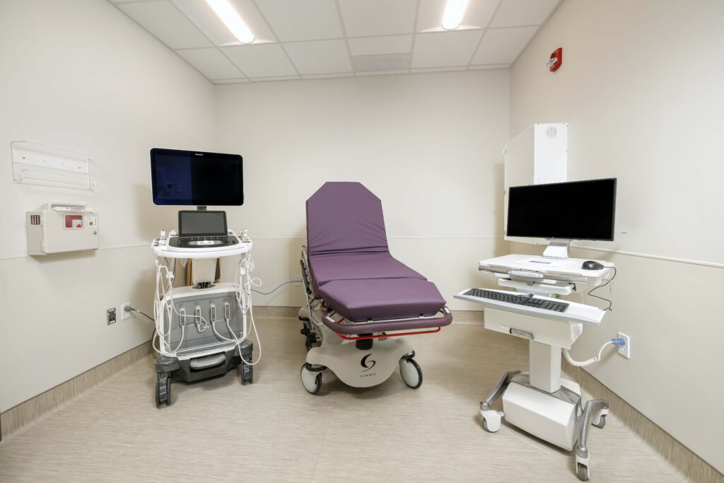 A medical examination room with an adjustable purple procedure table flanked by an ultrasound machine and a computer workstation.