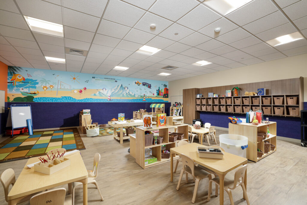 A preschool classroom with wooden tables, shelving units filled with toys, and a colorful mural featuring a quote about reading on the back wall.