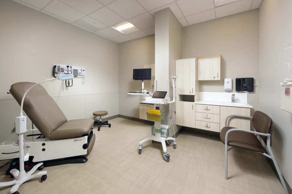 A medical examination room with an adjustable exam table, a rolling computer cart, cabinetry, and chairs.