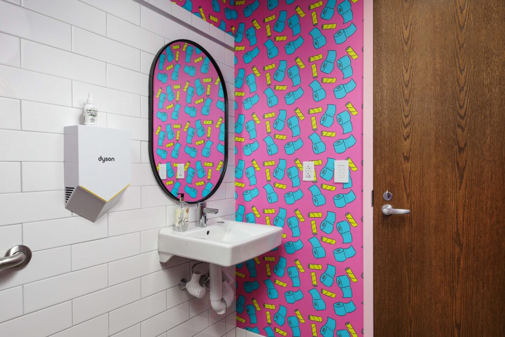 A bathroom sink and mirror on a wall with bright pink toilet paper patterned wallpaper, next to a white tile wall with a hand dryer.