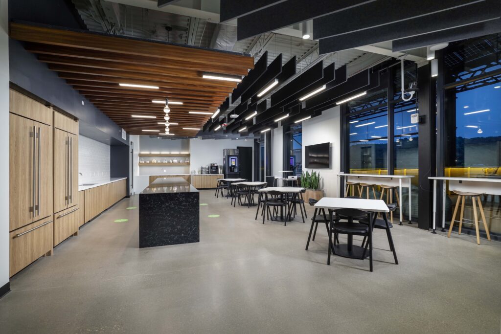 Modern office breakroom with a large kitchen island, wooden cabinetry, and several tables with chairs.