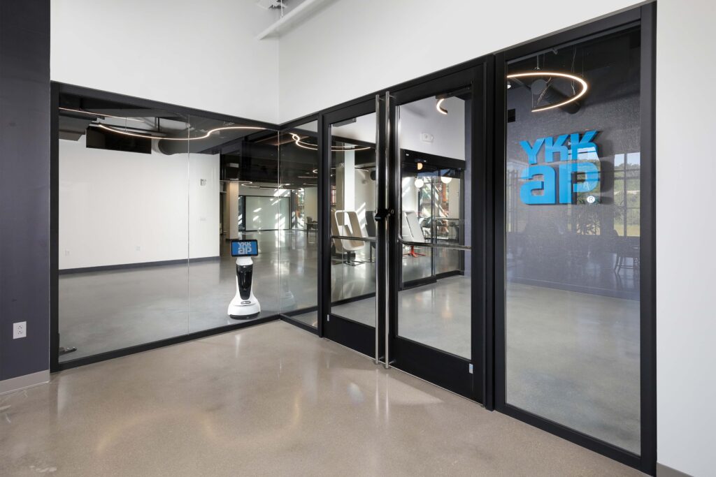 A small service robot stands in the lobby of an office space with YKK AP branding on a glass door.