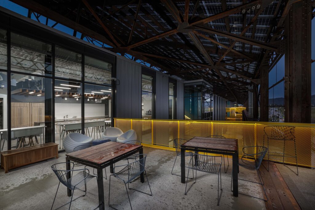 Outdoor patio with tables and chairs next to an office building at dusk, protected by a large metal roof structure with solar panels.