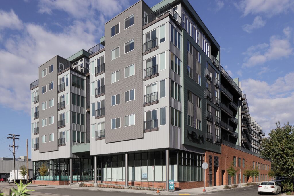 A multi-story modern apartment building featuring gray siding, balconies, and ground-floor storefronts with signage for “Helm on the Waterfront.”