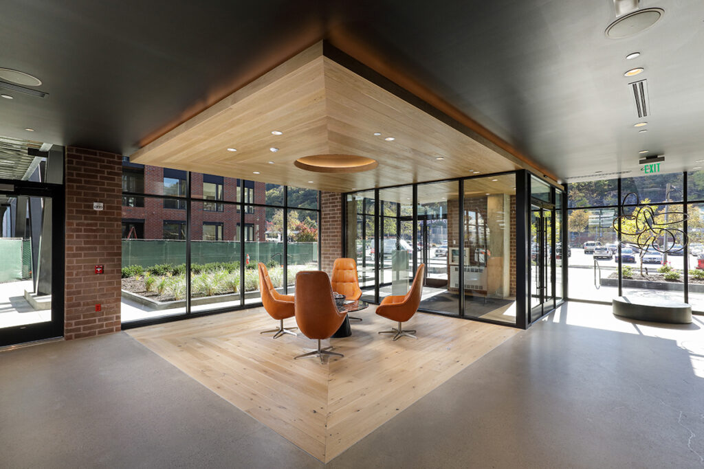A lounge area with four orange swivel chairs around a small table, situated under a wood-paneled ceiling feature in a building with large windows and brick walls.