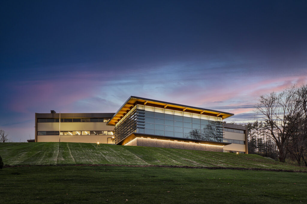 Modern multi-story office building with large glass windows and an overhanging roof, situated on a grassy hill at twilight.