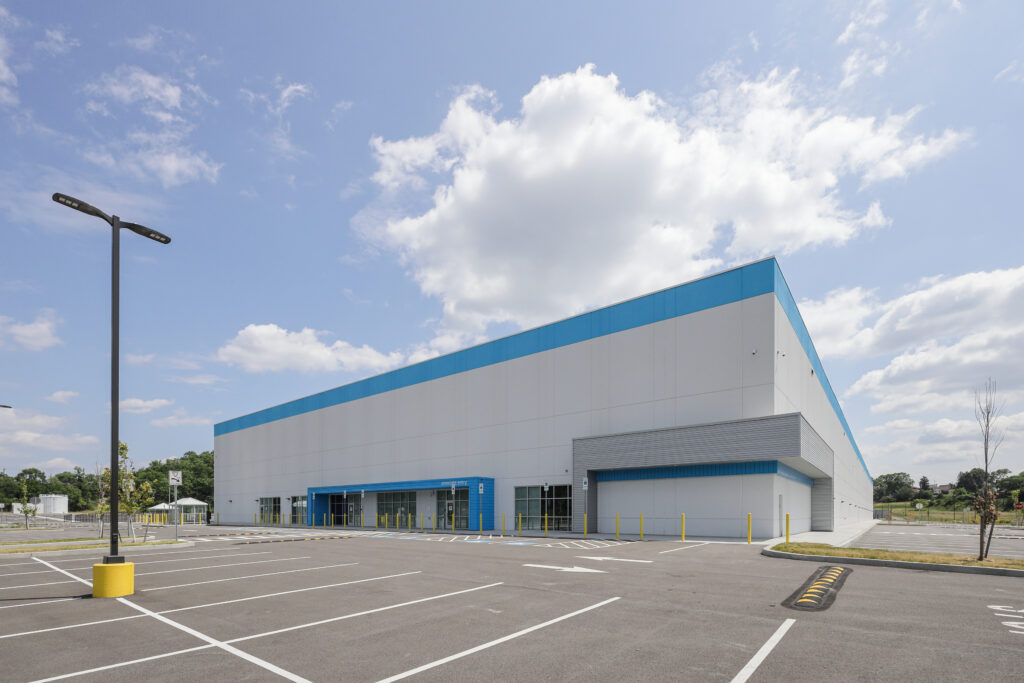 Exterior view of a large warehouse building with a white and blue facade, located in a spacious, mostly empty parking lot under a blue sky with clouds.