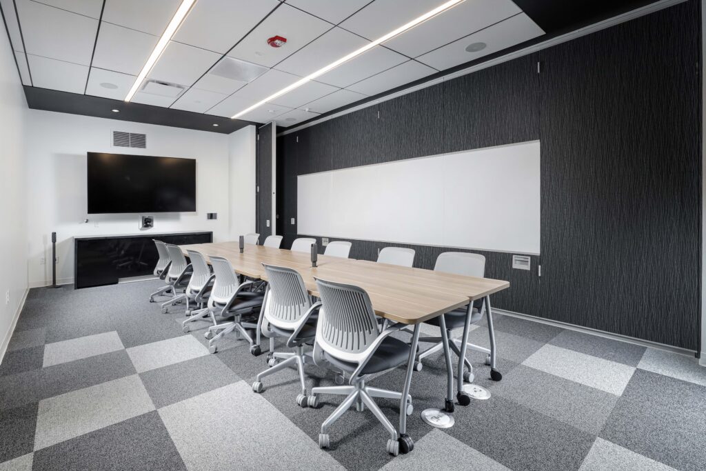 A modern conference room with a long wooden table, ergonomic chairs, a wall-mounted flat-screen television, and a large whiteboard.