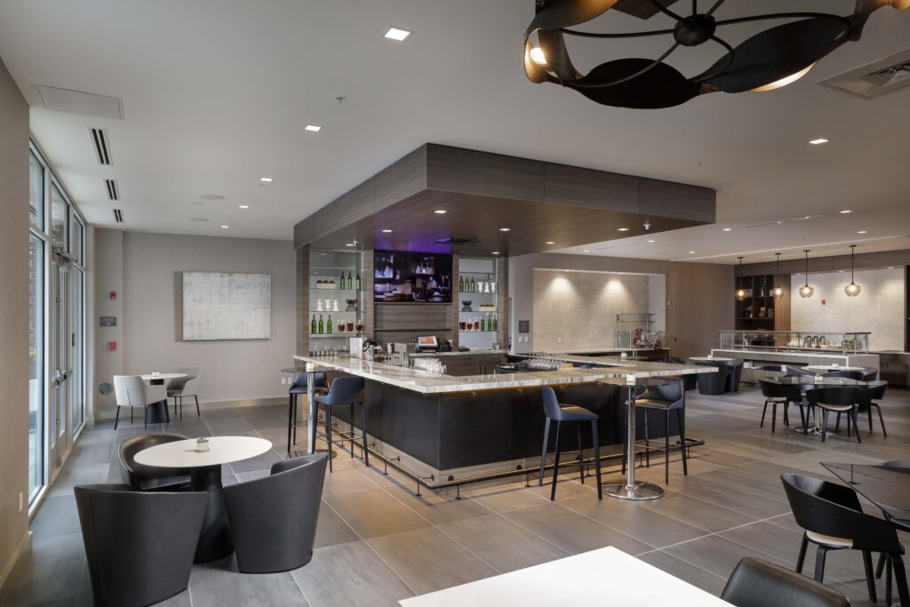 A restaurant interior featuring a central bar with stools, surrounding tables with chairs, and a television mounted above the back bar.