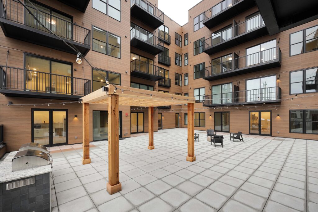 A modern apartment building courtyard with a wood pergola, a stainless steel grill, and small black patio chairs on a paved terrace.