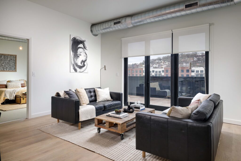 Living room with two black leather sofas facing a pallet coffee table, with a view of a city hillside through glass doors.