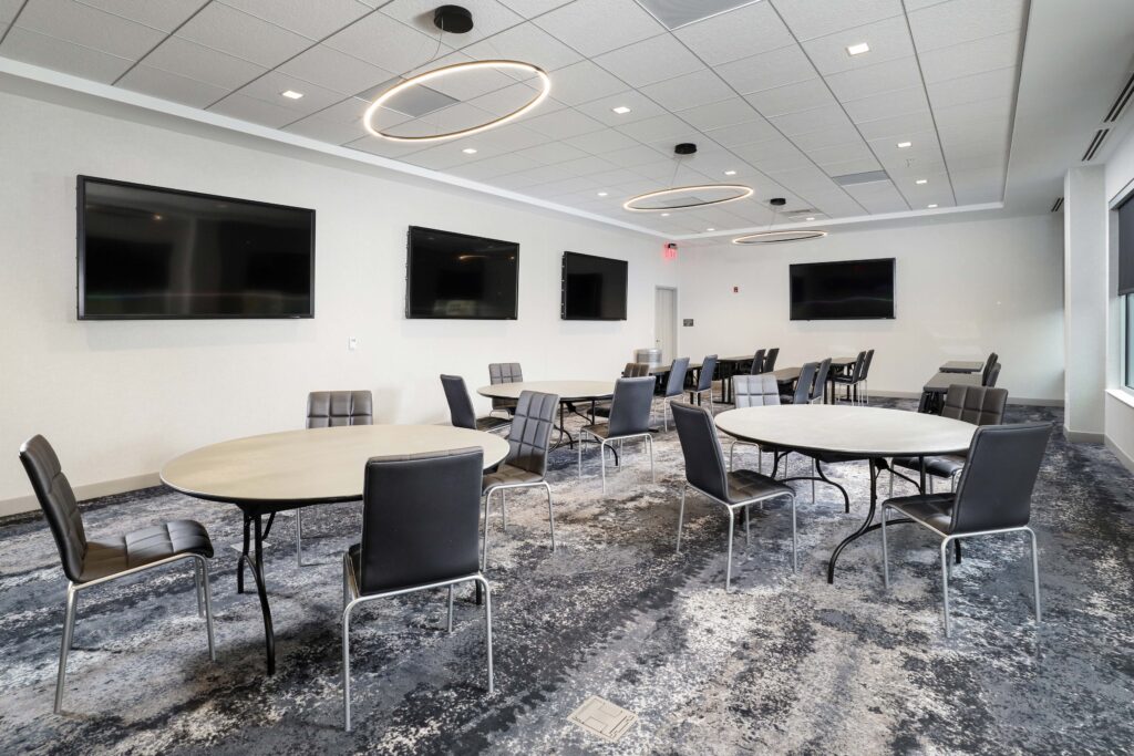 A conference room with multiple round tables and black chairs, featuring four flat-screen televisions mounted on the wall.