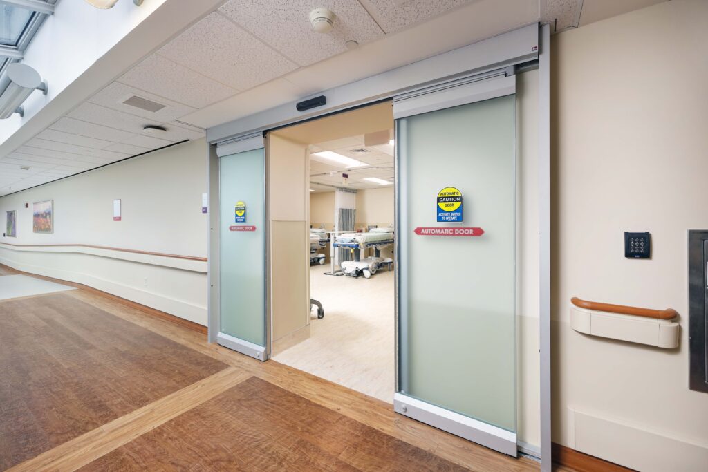 Open automatic sliding glass doors leading into a hospital room with a patient bed.