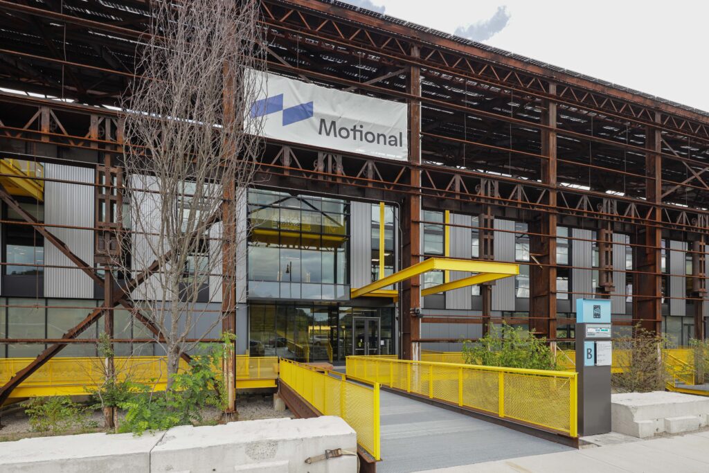 Exterior of a modern industrial-style office building with a banner reading “Motional” above the glass entrance and a yellow metal ramp leading to the door.