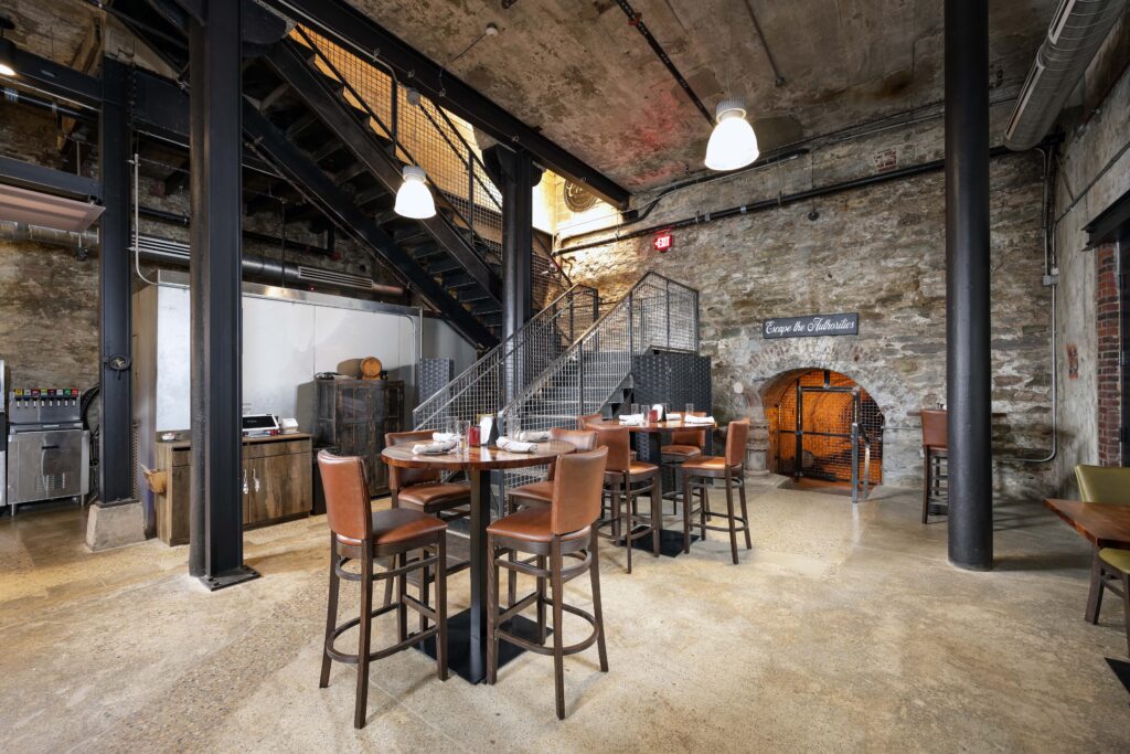 Dining area in a rustic, industrial space with a metal staircase, stone walls, and high-top tables with bar stools.