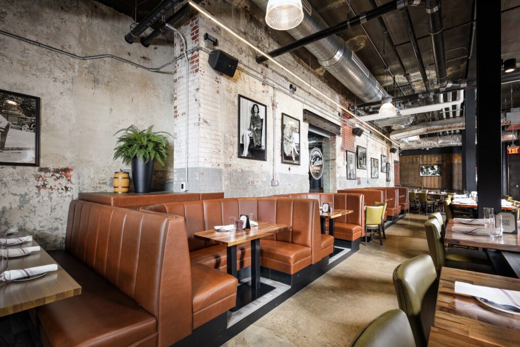 Dining room with brown leather booths, wooden tables, and exposed industrial ceiling pipes in a building with brick walls.